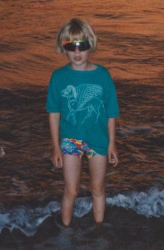 Rosie, aged 6, wearing a a terquoise t-shirt and sunglasses stands in the sea at sunset