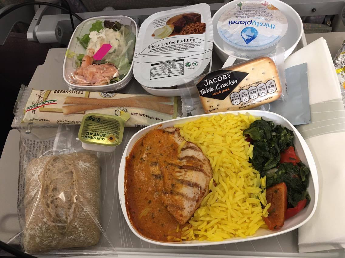 Emirates economy food with bread roll, sunflower spread, breadsticks, Jacob's Table Crackers, chicken curry with yellow rice and vegetables, a sticky toffee pudding and a thirstyplanet water on a grey tray