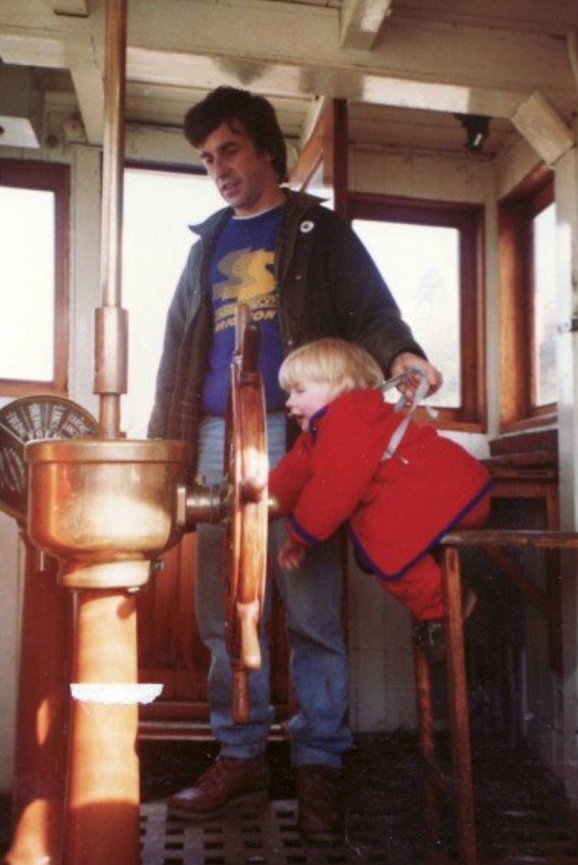 Rosie as a toddler, wearing a red coat and reins, held up by her dad, to a wooden boat wheel