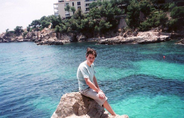A teenager wearing a t-shirt and shorts sits on a rock in front of a terquioise bay in Majorca