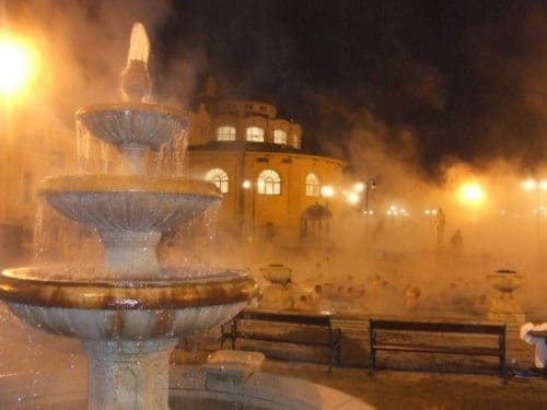 a 3 tiered water fountain in front of a large steaming pool at night at the Széchenyi gyógyfürdő Széchenyi Medicinal Bath in Városliget City Park, Budapest