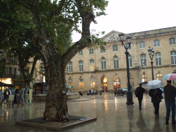 People with umbrellas walking through Place de l'Hôtel de Ville, with the Halle aux Grains in the background, Aix-en-Provence, France