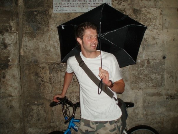 A young man in a white t-shirt holds a black umbrella and leans against his bike