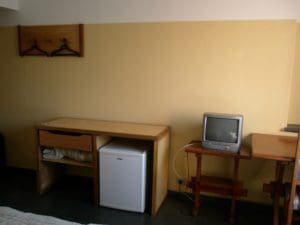 A yellow wall of a hostel room. A table and drawer with under counter fridge. A a grey TV on a stand. Hangers on hooks on higher on the wall.