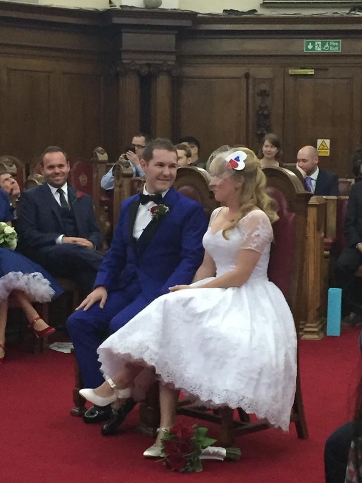 Karl in a blue 3 piece suit and Rosie in a white shin length dress sit surrounded by family and friends in the wood panelled Council Chamber, Islington Town Hall, London about to get married