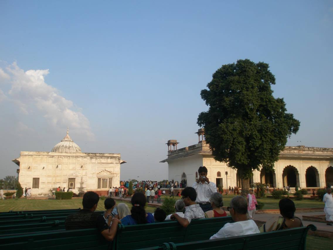 Indian tourists sitting on green benches in the gardens of Red Fort Delhi, India, in the background the white marble Khas Mahal