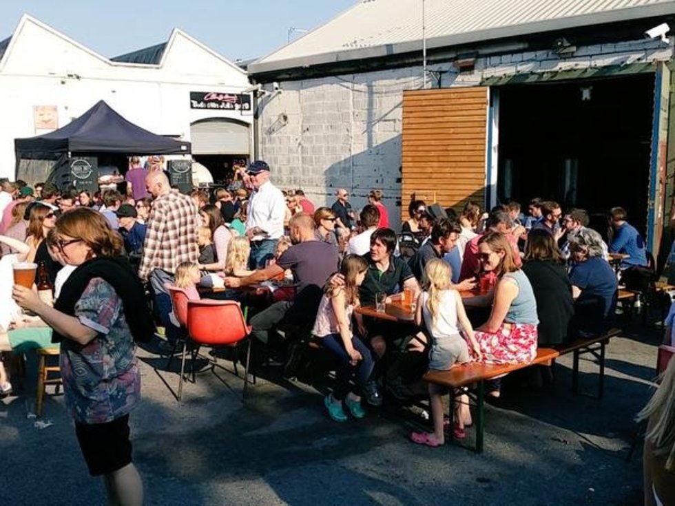 people sitting at tables in the sunshine, a white bricked building in the background is The Wildcard Brewery Barrell Store, Walthamstow, London