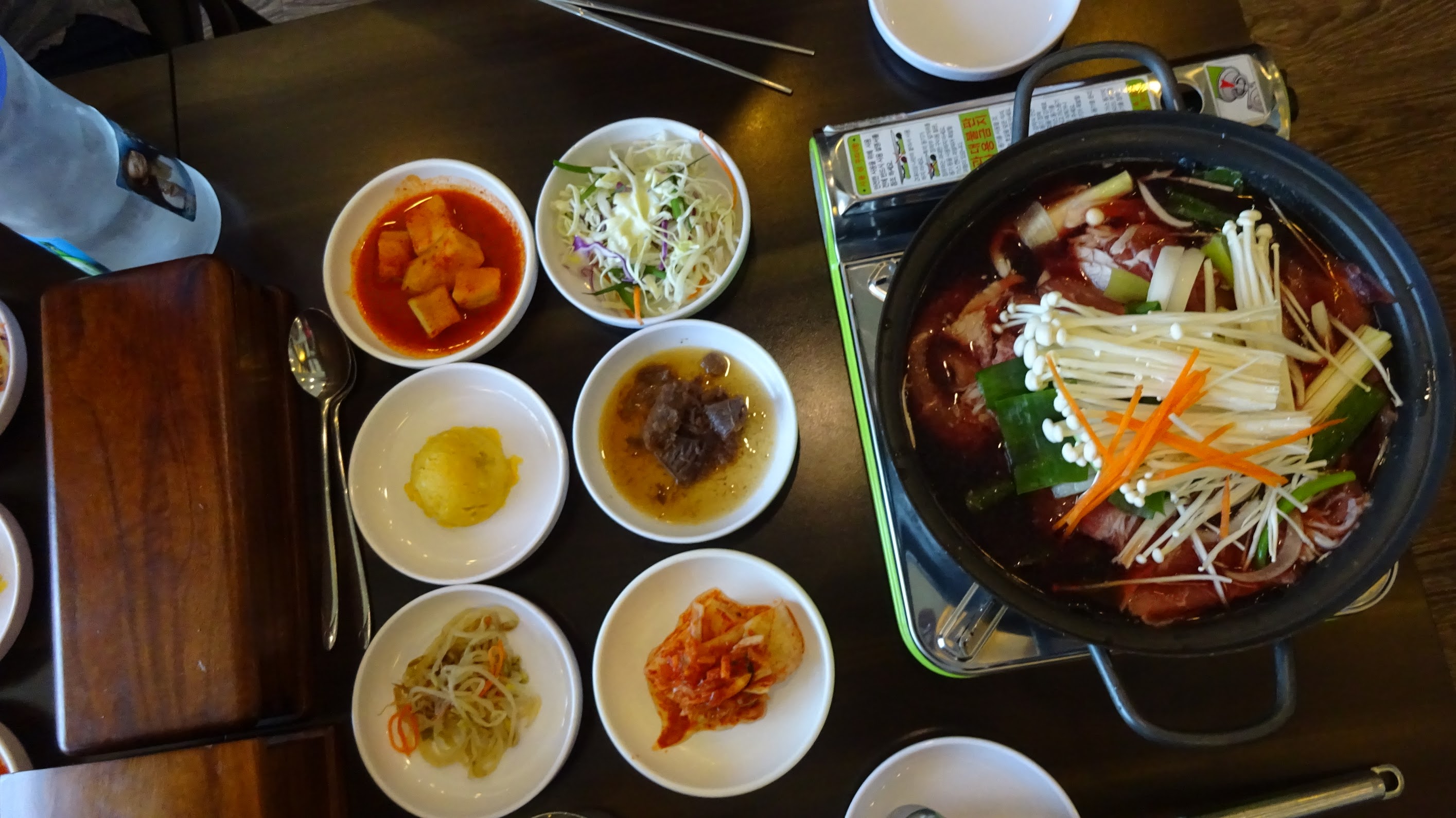 A black metal pot with beef bulgogi in a broth with vegetables on top. Next to it are 6 white bowls of side dishes at Imjingak Park, South Korea