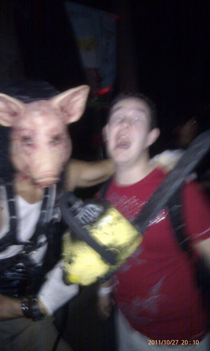 Karl wearing a red t-shirt, pretending to scream as a person in a pig mask, with his arm around Karl, holds a yellow chainsaw below Karl's neck