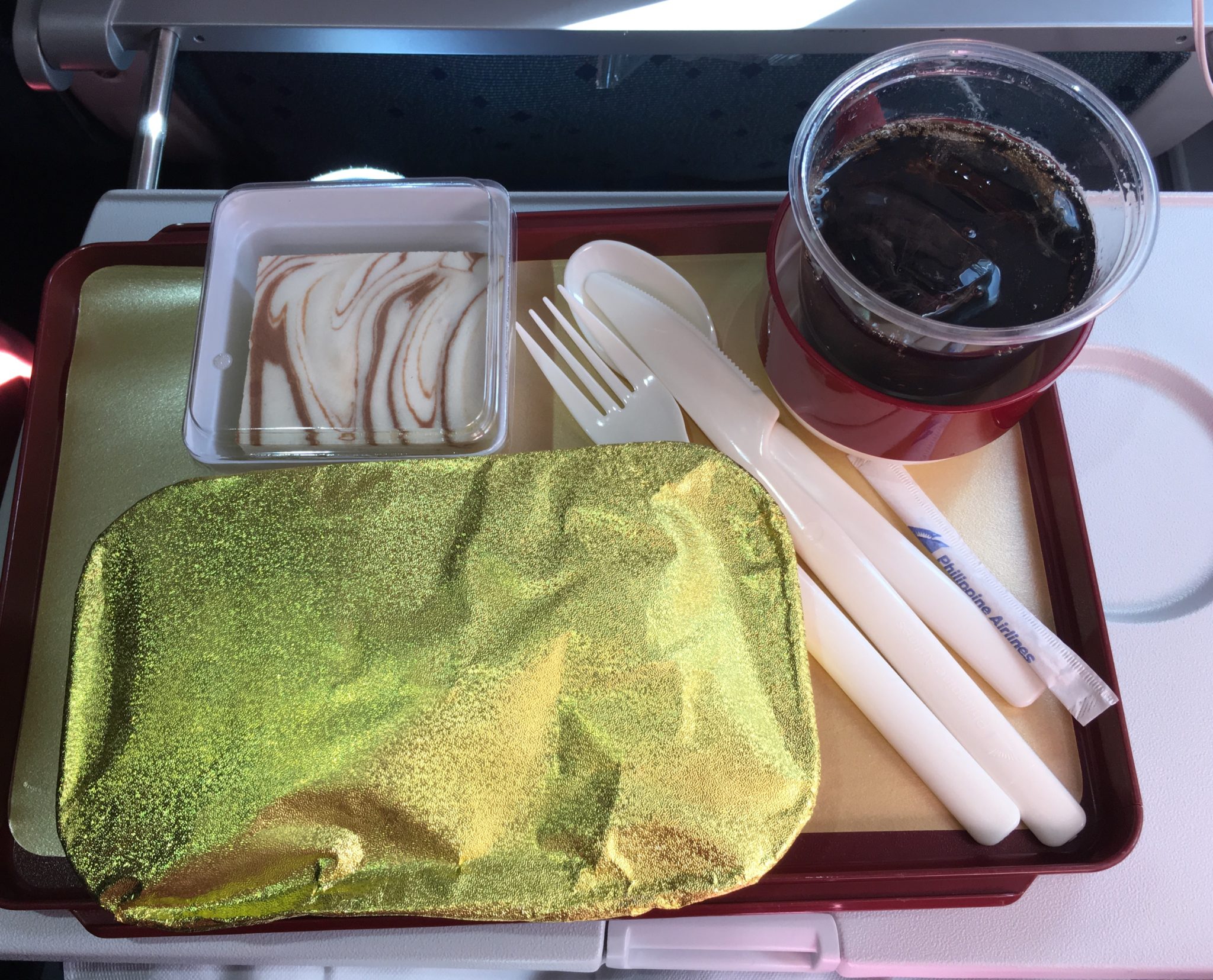 In flight meal on a red plastic tray with a gold foil tray, cream plastic cutlery, a square cheesecake and a plastic cup of Coke on economy Philippine Airlines A330-300 plane