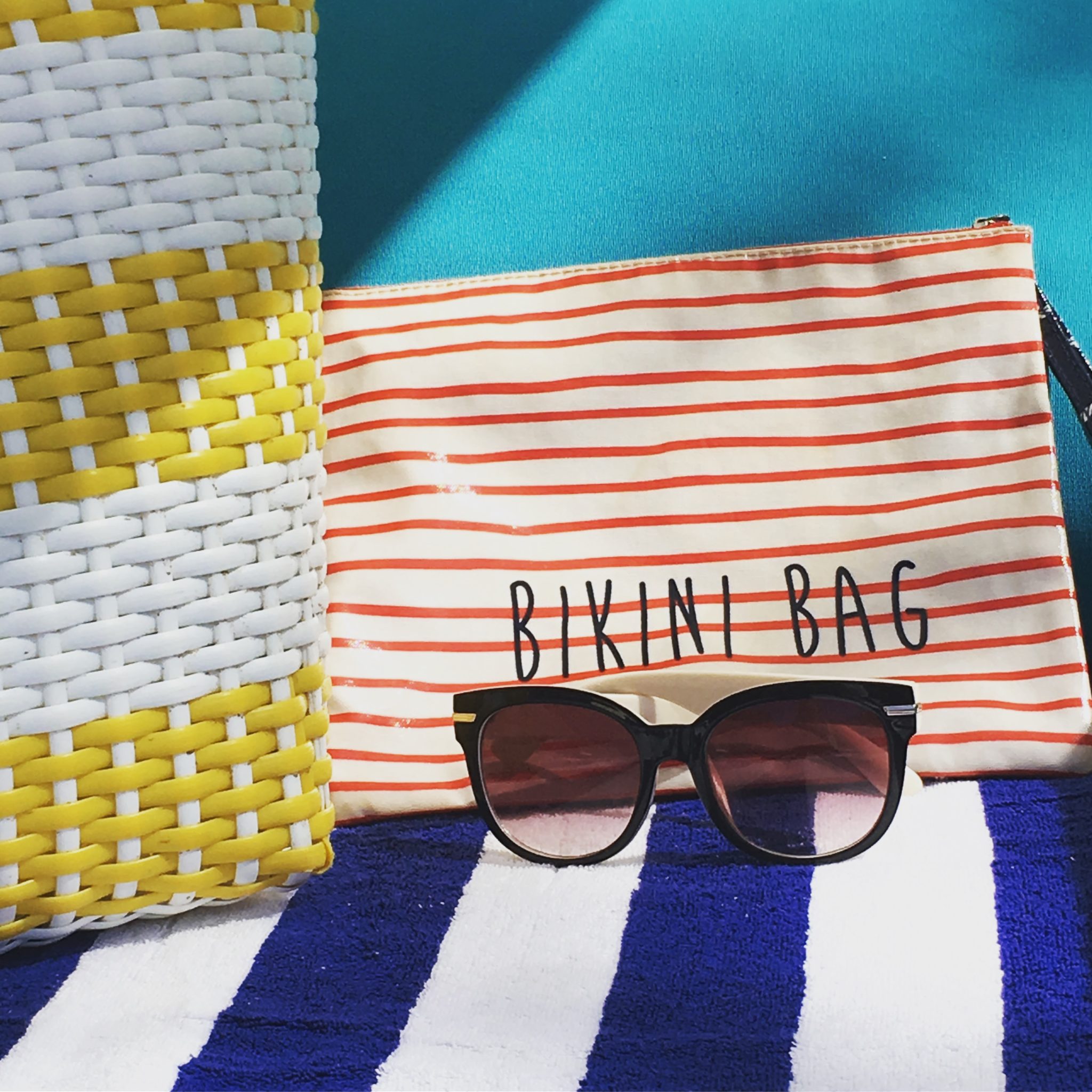 Pink sunglasses, striped bikini bag and white and yellow rattan bag on a blue and white striped towel