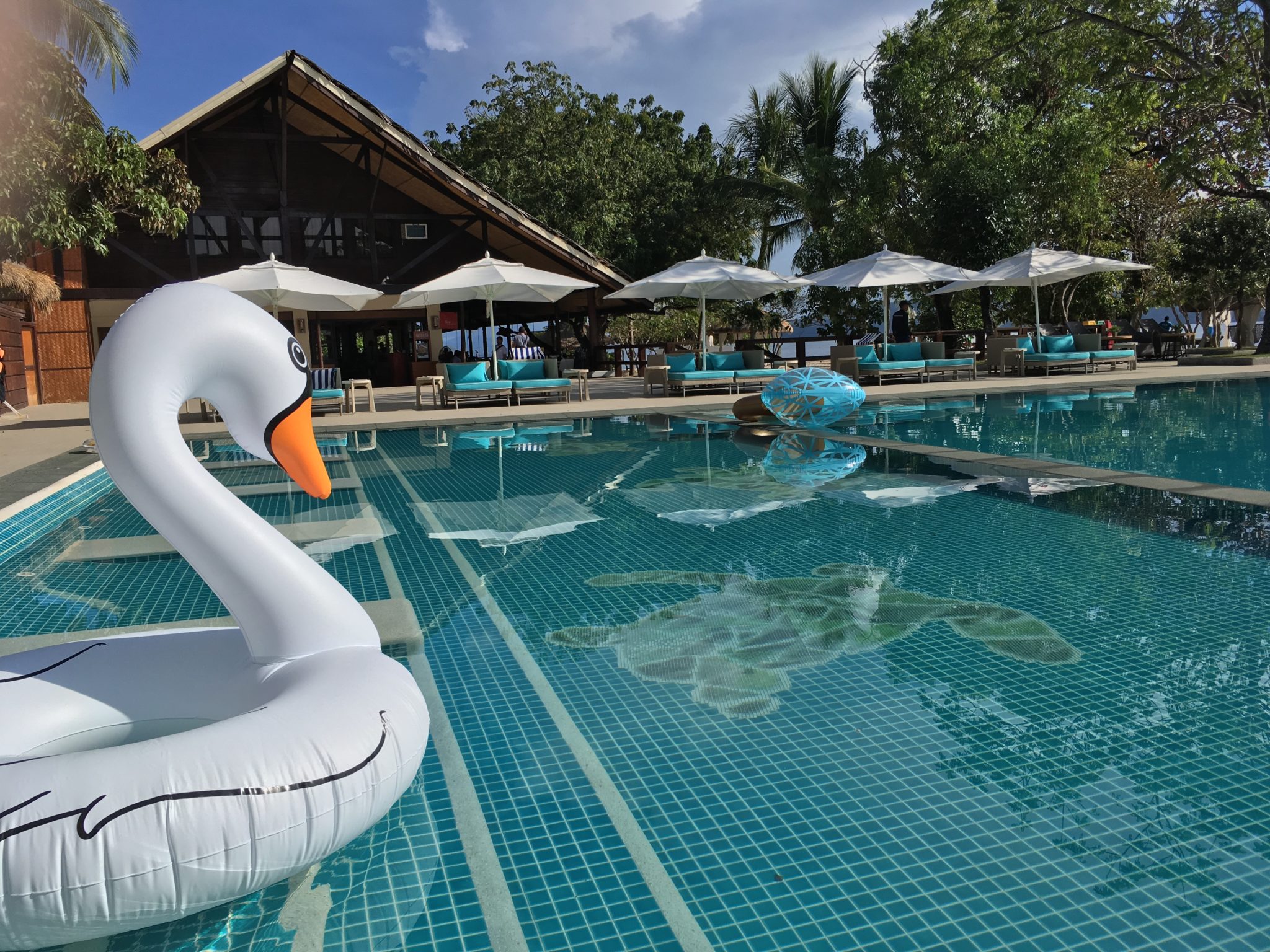 swimming pool with an inflatable swan. In the background blue sun loungers and white parasols in front of trees at Club Paradise Palawan