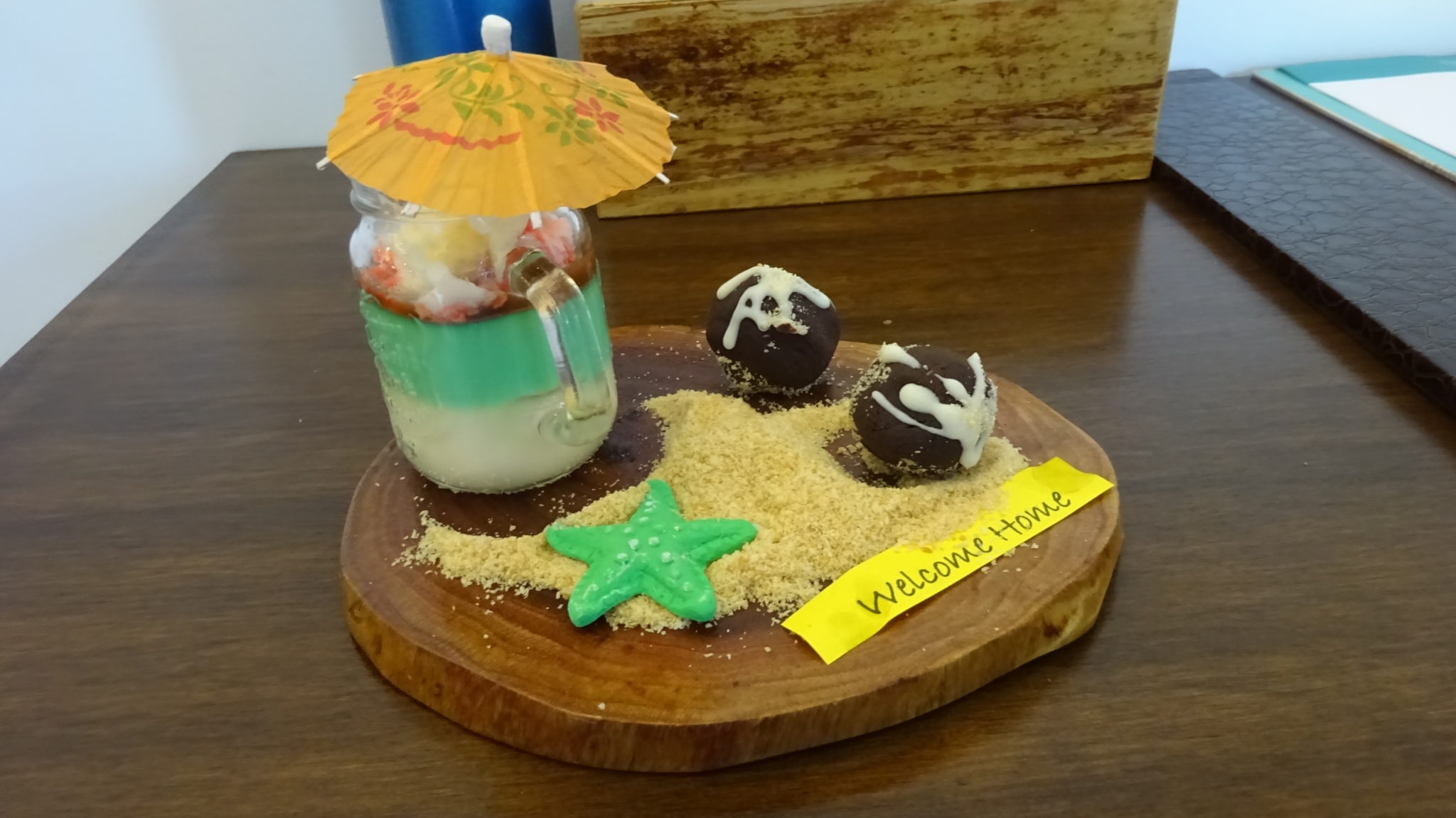 a panna cotta style dessert in a glass jar and 2 chocolate rum balls and sweet sand with 'Welcome Home' written on a piece of yellow paper