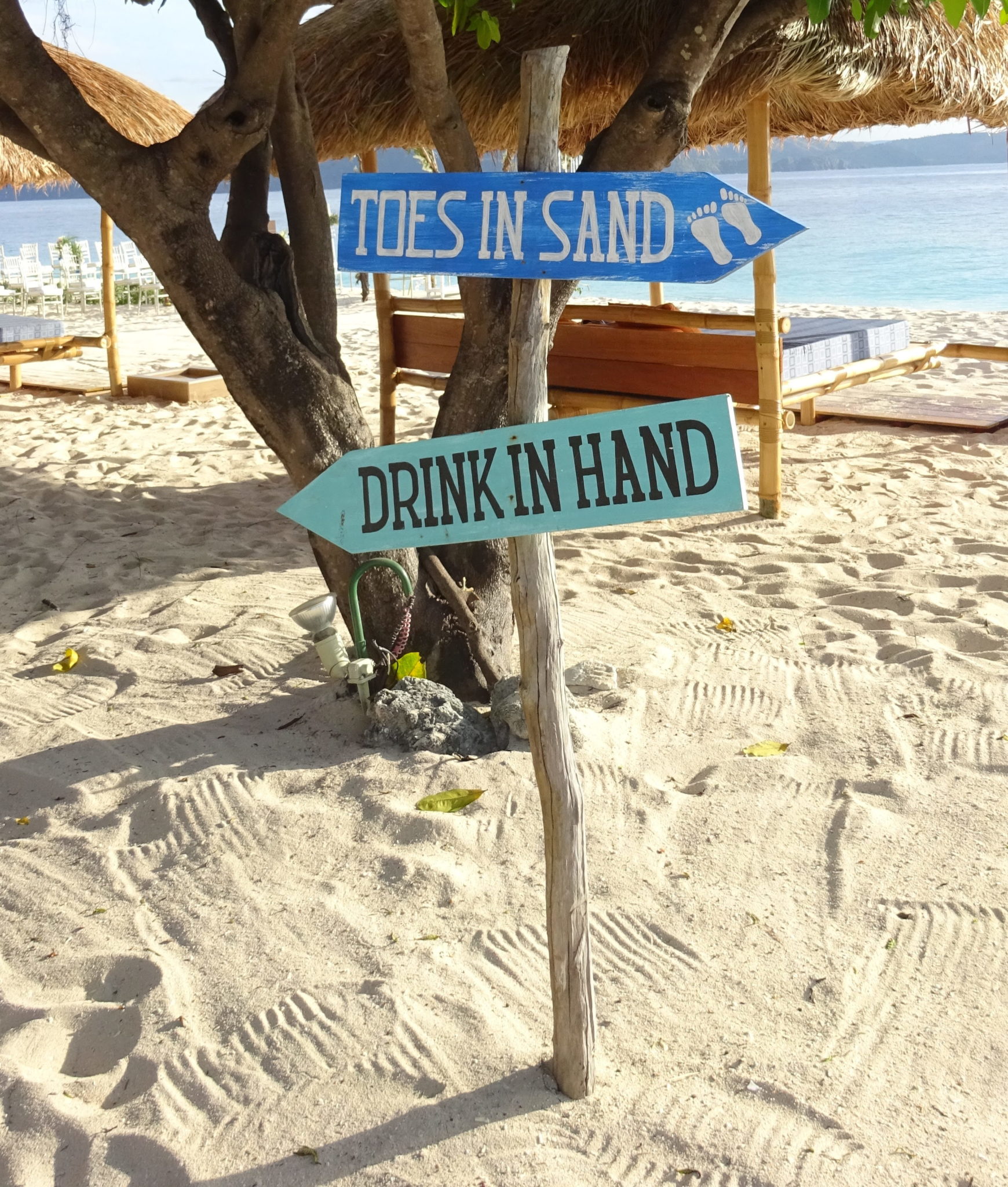 Wooden signs on a post in the beach reads 'toes in sand' and 'drink in hand'