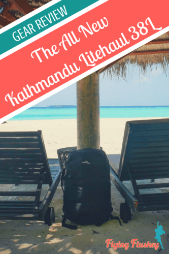 a black Kathmandu Litehaul 38L Backpack propped against a straw parasol on a white sandy beach in the Maldives. White text on a red banner in the top left corner reads 'The All New Kathmandu Litehaul 38L'