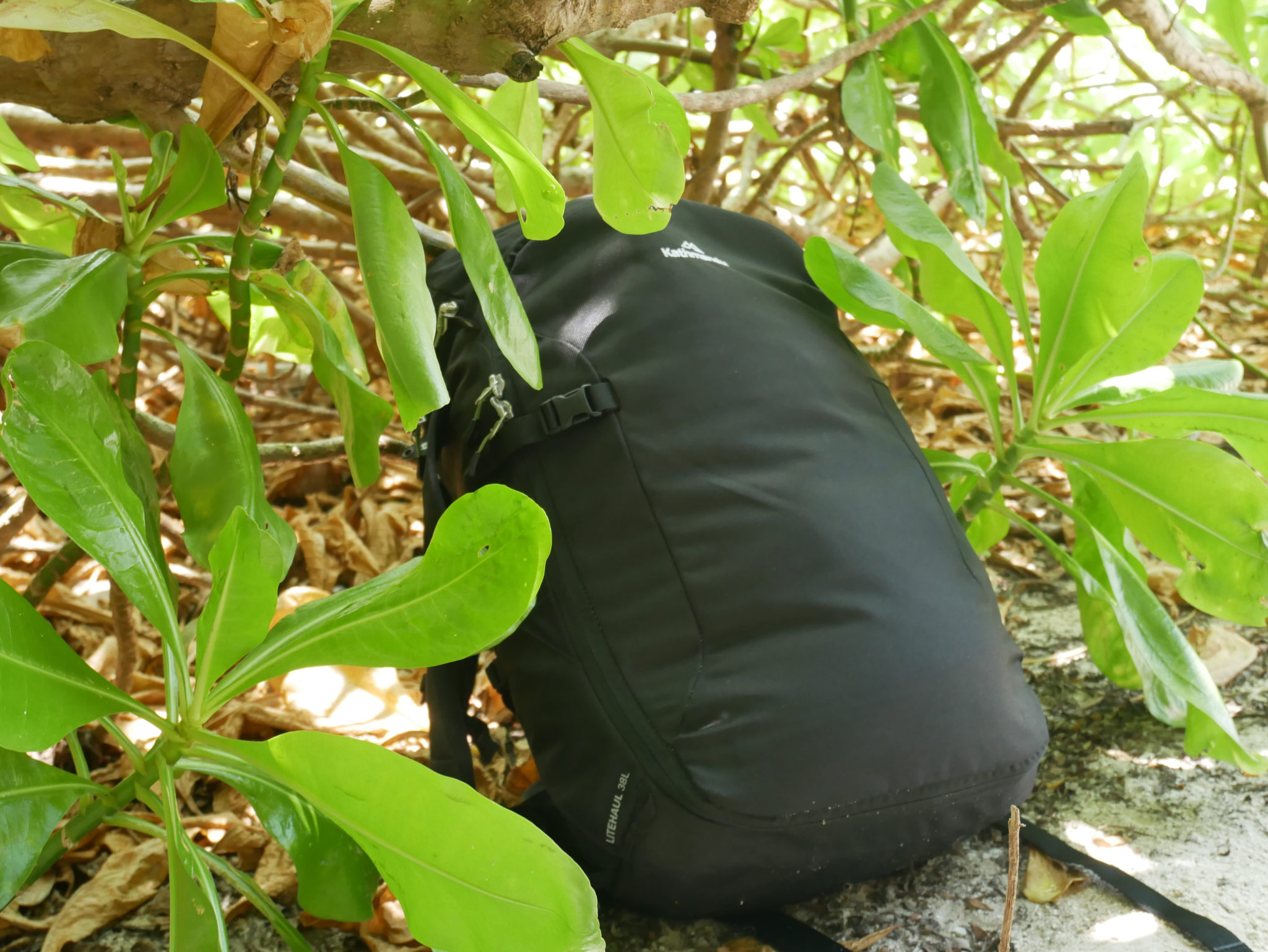 a black Kathmandu Litehaul 38L Backpack on the ground next to bushes in the Maldives