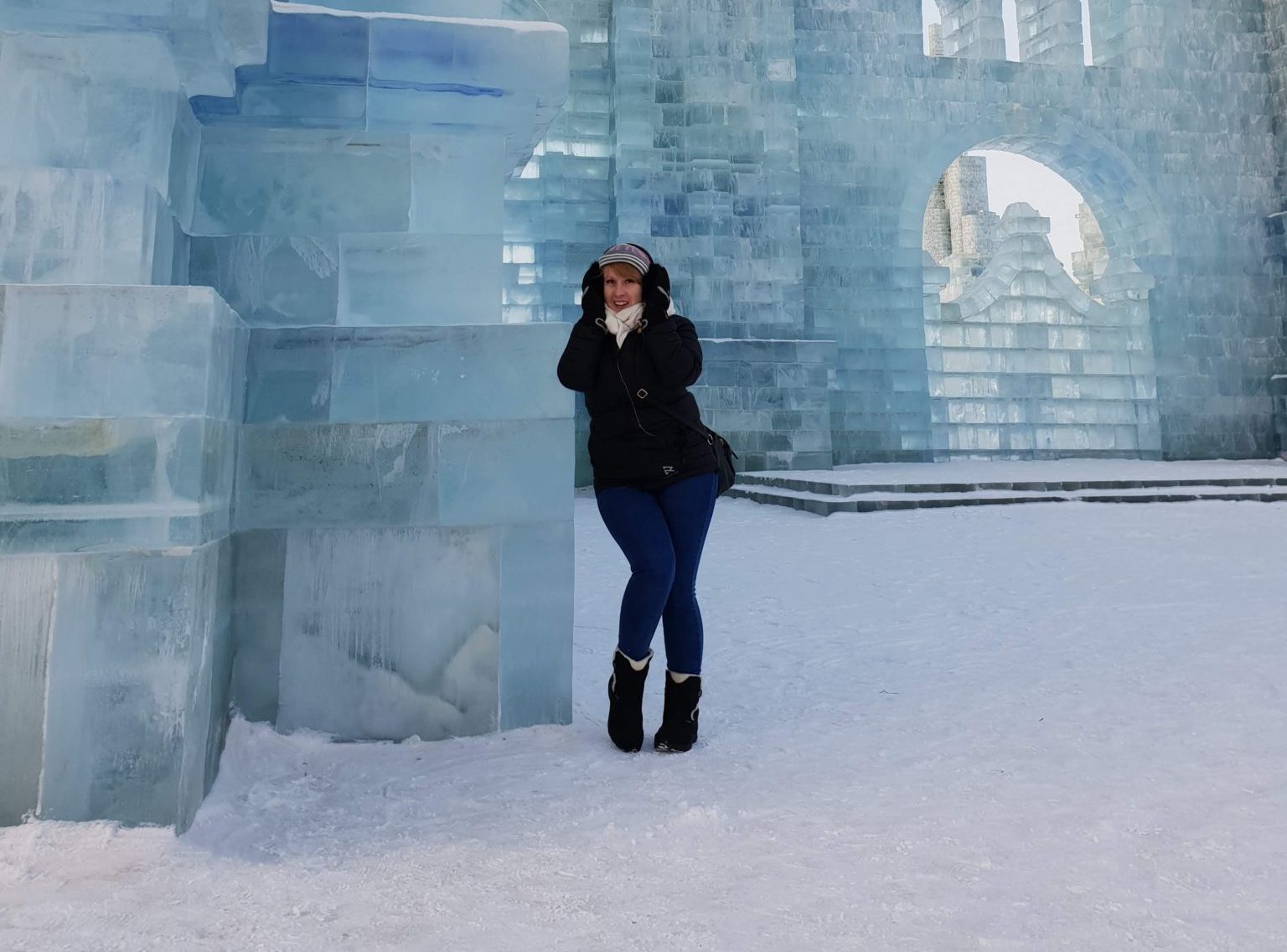 Rosie leans against an ice building wearing a coat and earmuffs at Harbin Snow and Ice Festival