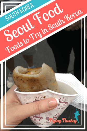 Bukkumi pan fried sweet potato tteok (rice cake), with a bite out of it, served takeaway in a smal paper coffee cup. Top left a blue and red diagonal banner with white text reads 'South Korea. Seoul Food. Foods to try in South Korea'