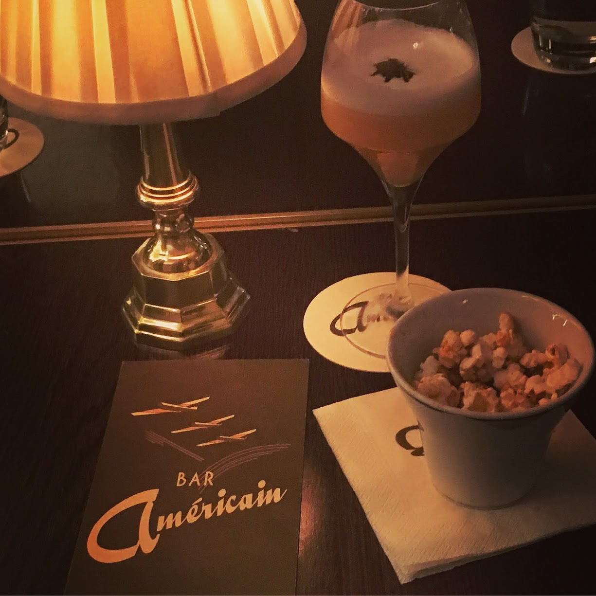 A silver table lamp, a wine glass with a foamy cocktail, a bowl of popcorn and a black menu card with Bar Americain in gold writing in Soho, London