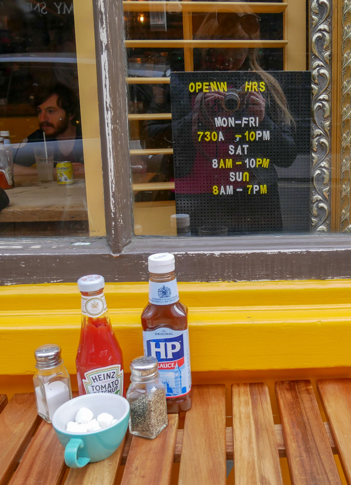 A wooden table with sauces, salt, pepper and cup of sugar cubes in front of an opening hours sign at The Breakfast Club, Soho, London
