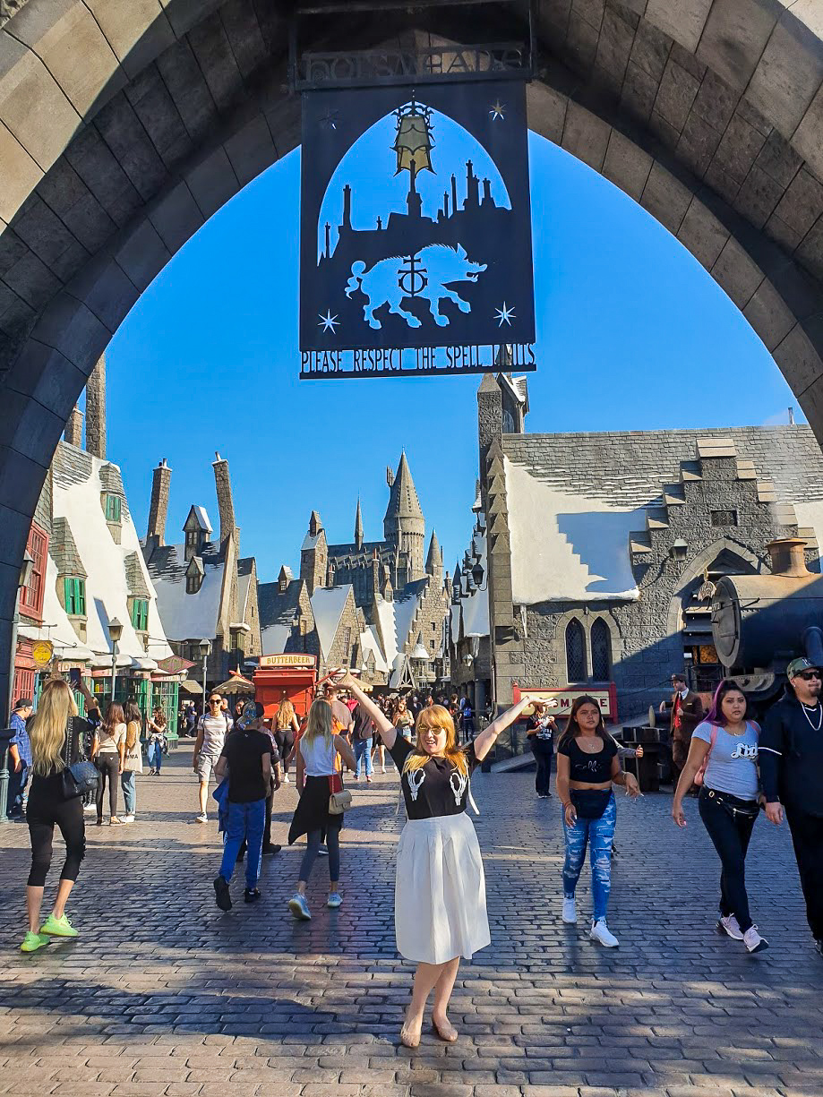 Rosie poses with her arms aloft for a photo beneath the Hogsmeade sign. Behind her, the roofs of Hogsmeade glisten with fake snow at Universal Studios Hollywood
