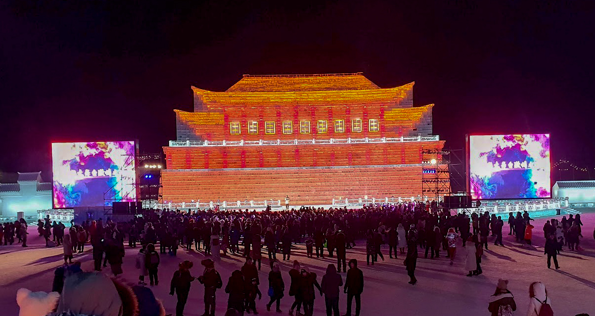 ice sculpture of traditional Chinese architecture, lit in red and oragne with a large screen either side at Harbin Ice and Snow World