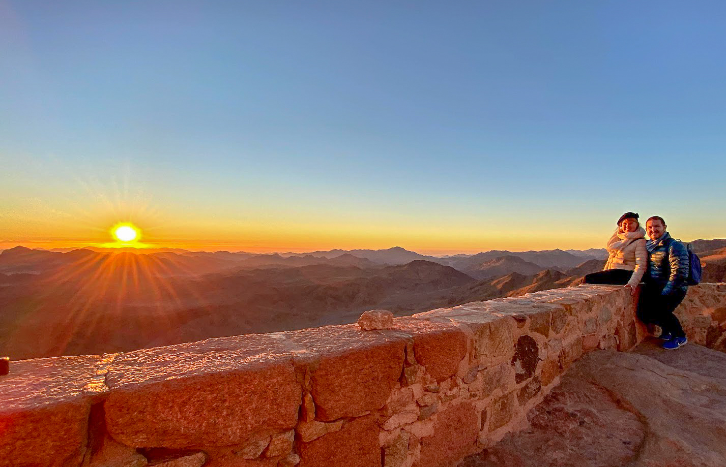 Rosie and Karl sitting on a wall as the sun rises at the summit of Mount Sinai, Egypt