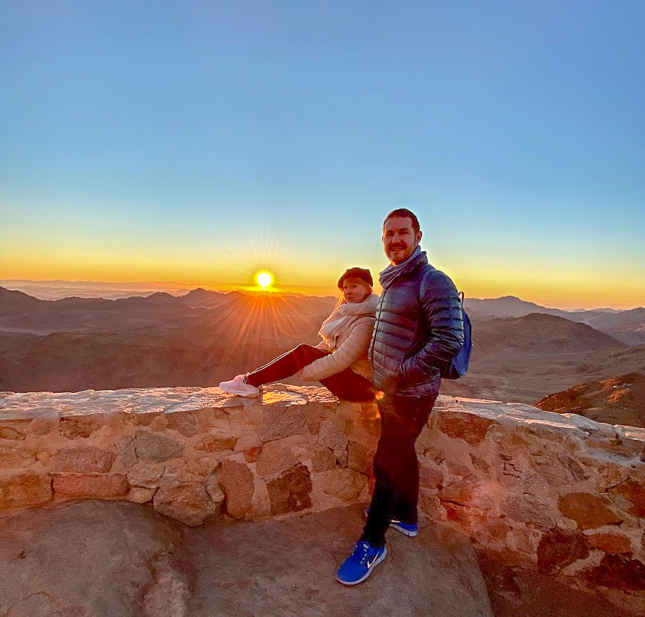 Rosie sitting on a wall beside Karl, standing, as the sun rises at the summit of Mount Sinai, Egypt