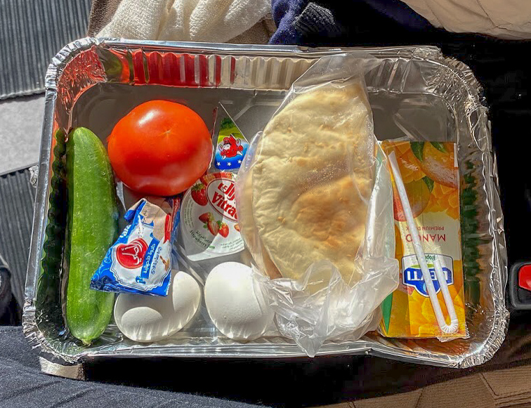 a foil tray with a whole cucumber, tomato, 2 white hard boiled eggs, some bread and a mango juice carton