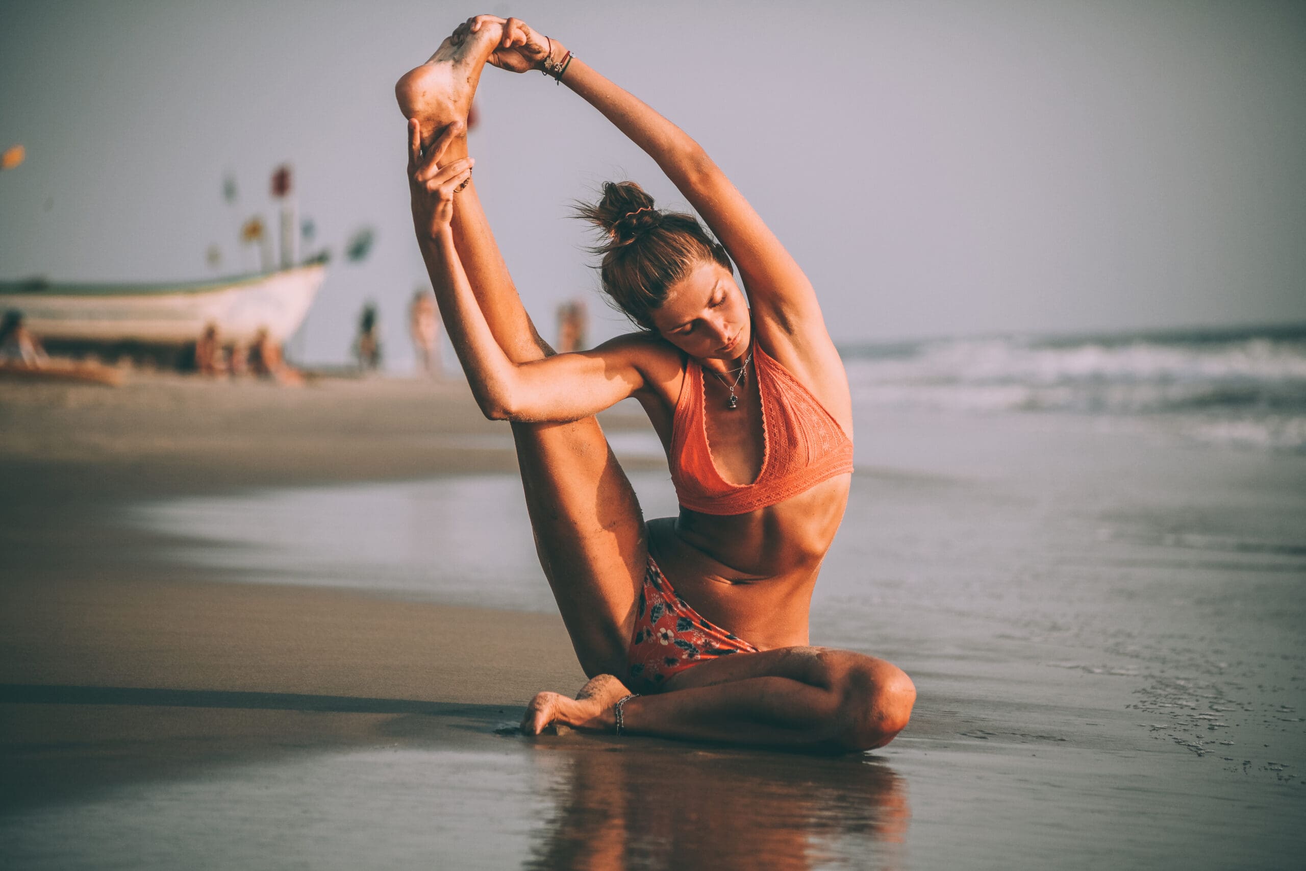 A woman in a peach bikini sitting on the wet sand against the sea stretches her right leg above her head in a yoga pose