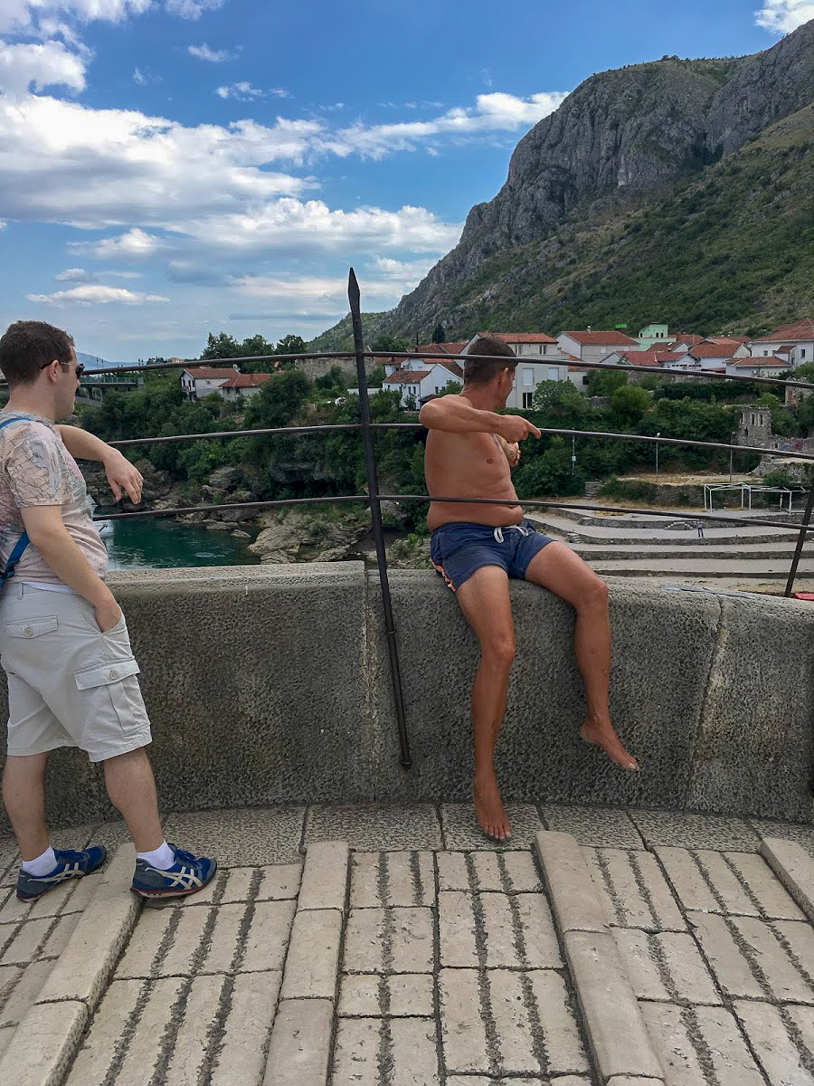 A man in swimming trunks sits on the edge of Stari Most Bridge, with Hum Hill in the background, Mostar