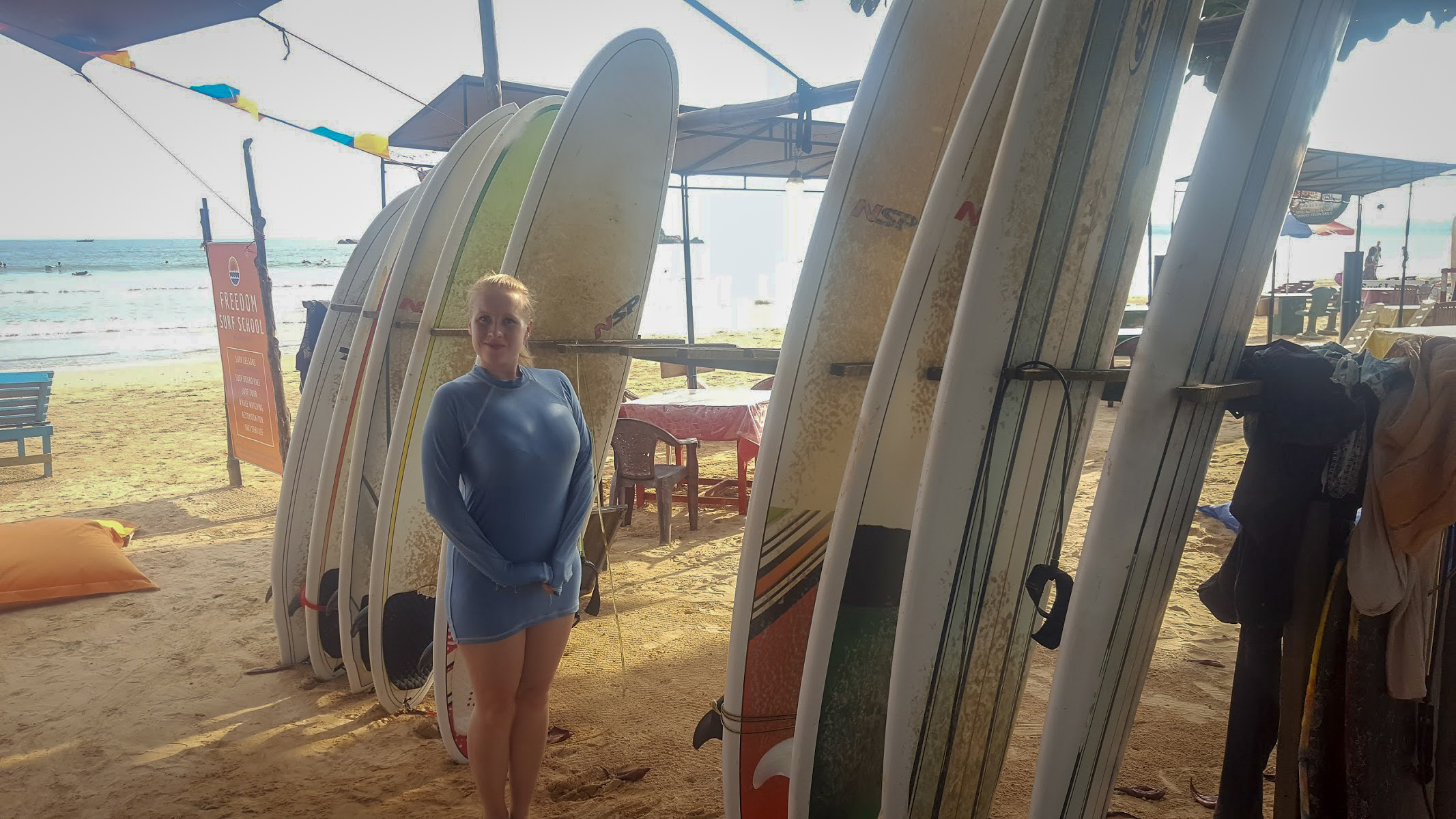 Rosie wearing a blue wet-shirt stands in front of surfboards on the beach in Sri Lanka