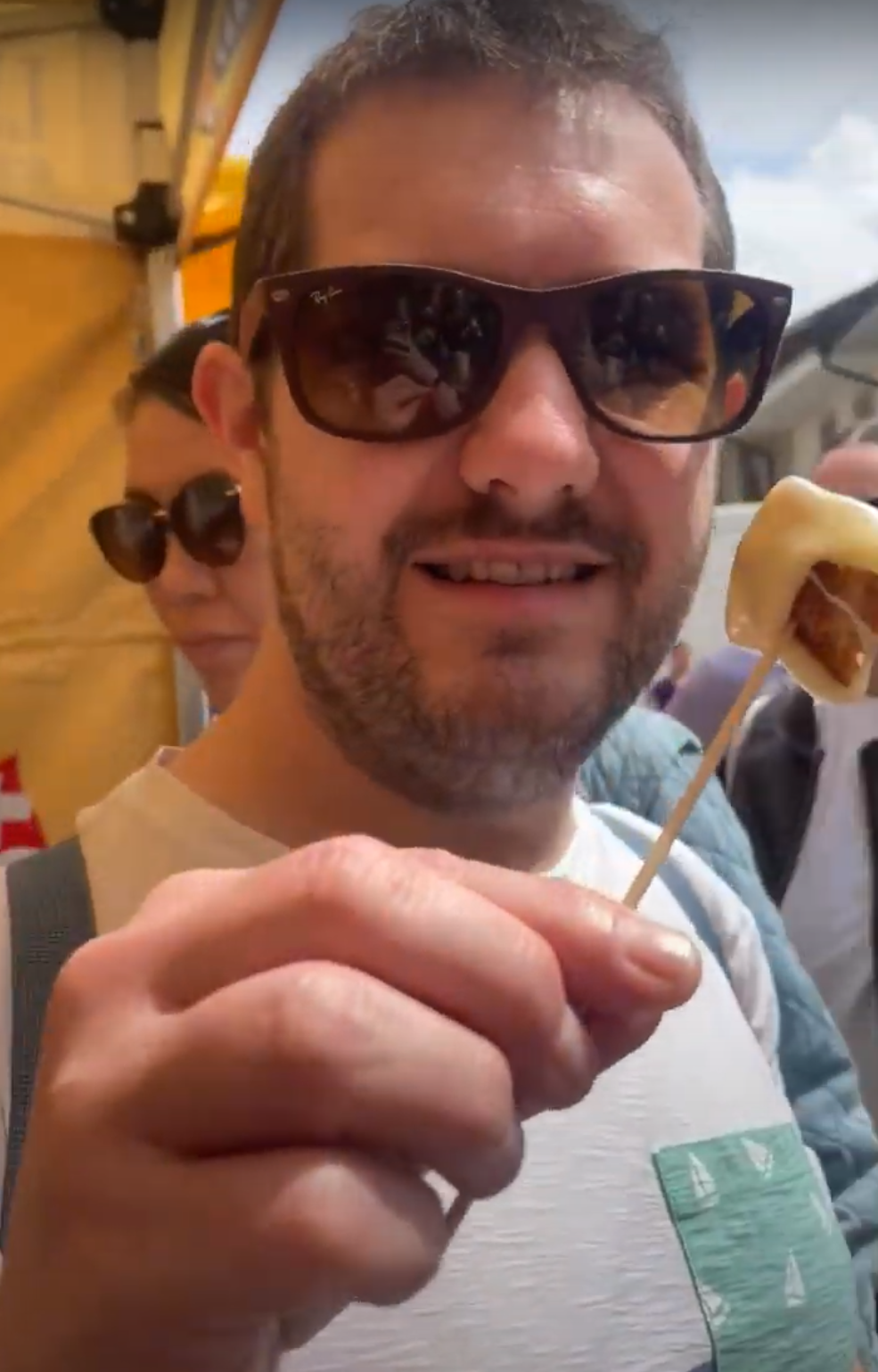 Karl wearing a sunglasses holds a stick with a bread covered in melted Gruyeres cheese